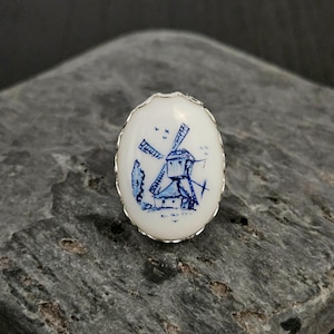 May include: Oval-shaped white and blue ring featuring a windmill design. The ring has a silver-colored, textured border. The windmill is depicted in blue, with a detailed illustration of the structure and surrounding trees.