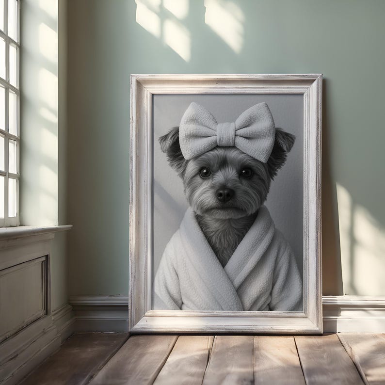 May include: A framed black and white portrait of a small dog wearing a white bow and a white robe. The dog has dark fur and is looking directly at the viewer. The frame is white and the background is a light green.