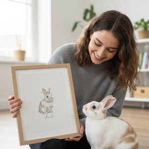 May include: A framed watercolor print of a rabbit with tan and white fur. The print is in a light wood frame. A live rabbit with similar coloring is next to the framed print. The word "Rosita" is written below the rabbit in the print.
