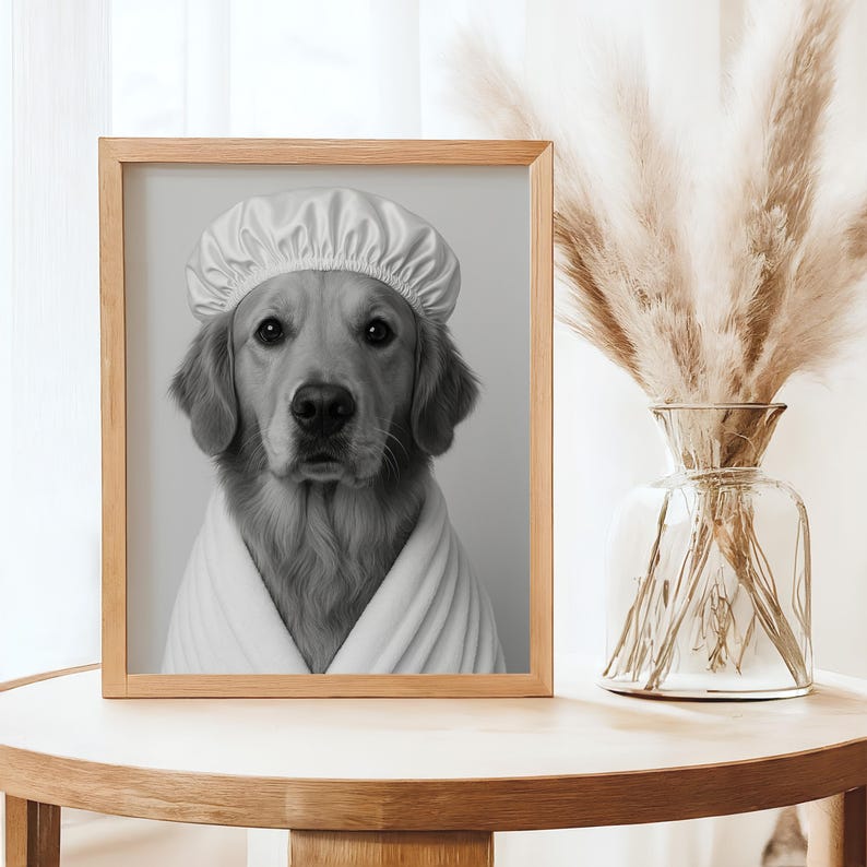May include: Black and white framed print of a Golden Retriever wearing a white shower cap and a white robe. The dog is looking directly at the viewer. The print is displayed on a wooden table next to a glass vase with dried pampas grass.