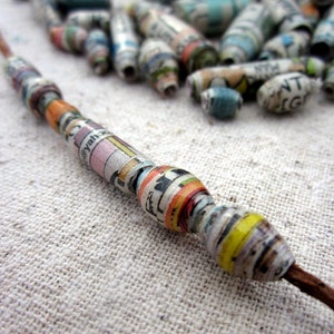 May include: A close-up of a necklace made of colorful paper beads. The beads are rolled from newspaper and magazine pages, creating a unique and textured look. The beads are strung on a brown cord.