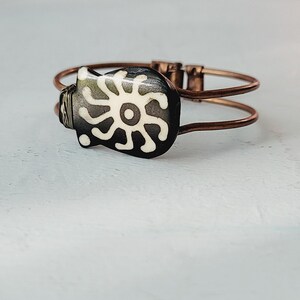 Könnte beinhalten: Ein Armband aus Kupferdraht mit einem schwarzen und weißen Keramik-Anhänger, der ein stilisiertes Sonnenmotiv zeigt.