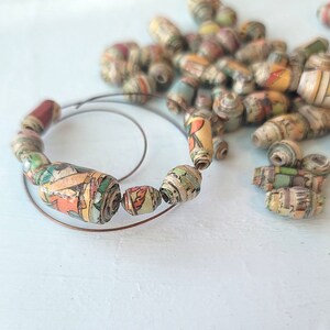 May include: A close-up of a necklace made of colorful paper beads. The beads are rolled from recycled comic book pages and strung on a copper wire. The necklace is lying on a white surface.
