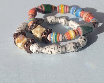 May include: Two bracelets made of rolled paper beads. One bracelet is white with brown accents and the other is multi-colored with green, orange, yellow, blue, and red beads.