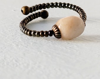 May include: A delicate ring made of small brown beads with a single large, oval-shaped, light pink stone in the center.