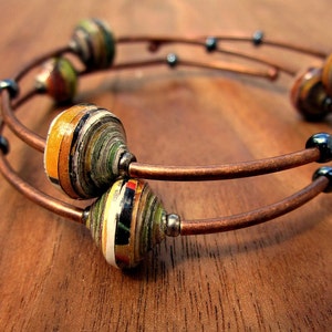 Könnte beinhalten: Ein doppelsträngiges Kupferdrahtarmband mit bunten Papierperlen. Die Perlen bestehen aus aufgerolltetem Papier und haben eine Vielzahl von Farben, darunter Gelb, Grün und Braun. Das Armband ist eine großartige Möglichkeit, jedem Outfit einen Hauch von Farbe und Stil zu verleihen.