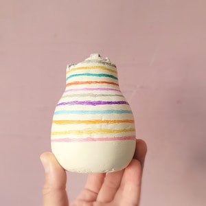 May include: A small, white ceramic vase with a striped pattern in various colors, including pink, yellow, orange, blue, purple, and gray.