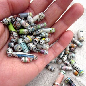 May include: A hand holding a collection of colorful paper beads made from rolled up magazine pages. The beads are various lengths and have different patterns and colors.
