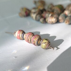 May include: A close-up of a string of beads made from recycled paper. The beads are in shades of pink, green, and brown, and have a textured surface. The beads are strung on a thin wire.