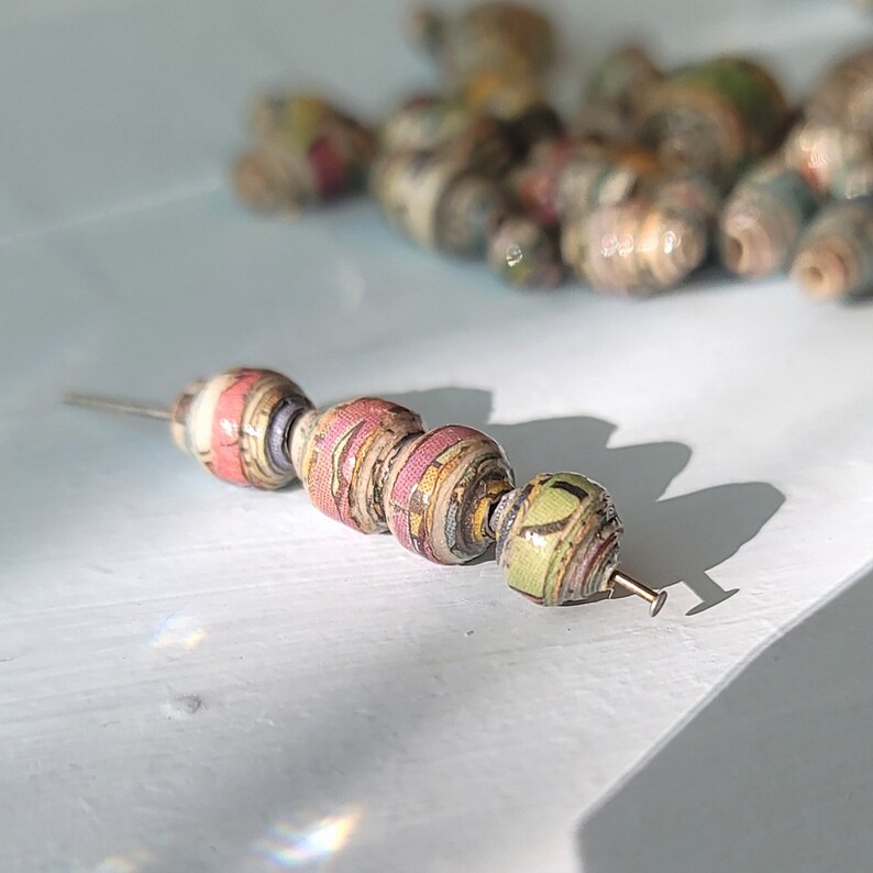 May include: A close-up of a string of beads made from recycled paper. The beads are in shades of pink, green, and brown, and have a textured surface. The beads are strung on a thin wire.