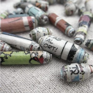 May include: A close-up of colorful, rolled paper beads. The beads are made from recycled paper and have a variety of patterns and colors. Some of the beads have text printed on them, including the words "AS AL ORT!"