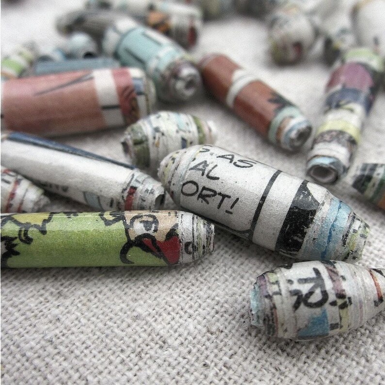 May include: A close-up of colorful, rolled paper beads. The beads are made from recycled paper and have a variety of patterns and colors. Some of the beads have text printed on them, including the words "AS AL ORT!"