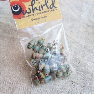May include: A clear plastic bag filled with colorful, handmade recycled paper beads. The bag has a label that reads "whirl handmade recycled paper beads by Ren&eacute;e Parker".