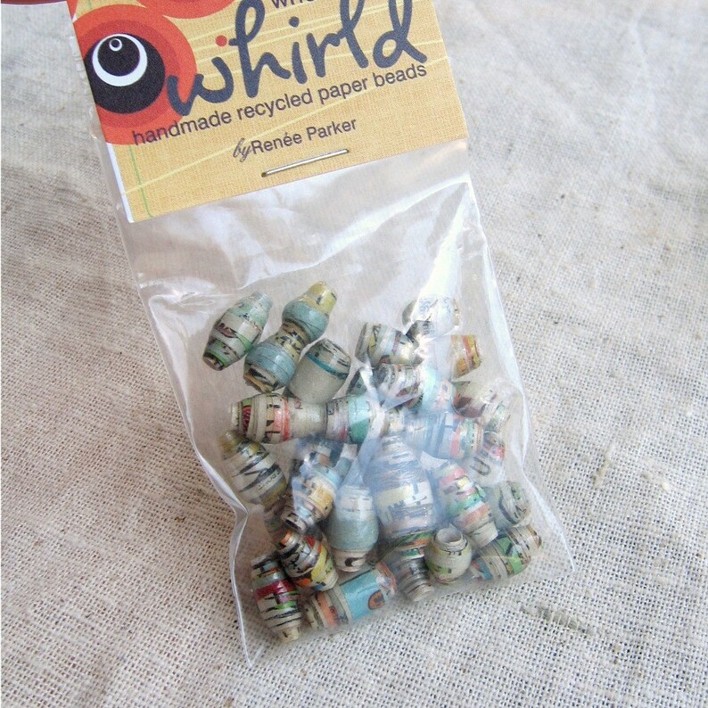 May include: A clear plastic bag filled with colorful, handmade recycled paper beads. The bag has a label that reads "whirl handmade recycled paper beads by Ren&eacute;e Parker".