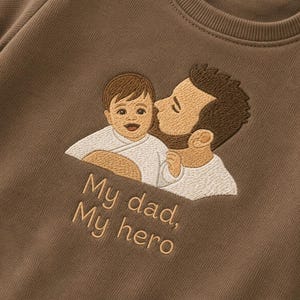 Op de afbeelding: Bruin sweatshirt met een geborduurd ontwerp van een vader die een baby kust. De tekst "My dad, My hero" is eronder geborduurd in een bijpassende kleur. De sweatshirt is effen van kleur.