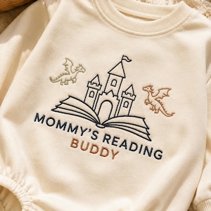 Può includere: Tutina per neonati color crema con design ricamato. Presenta un castello, draghi e la scritta "MOMMY'S READING BUDDY" in filo blu navy e marrone. La tutina ha maniche lunghe e scollatura rotonda.