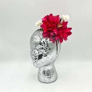 May include: A silver disco ball head with a vibrant fuchsia flower and small white flowers attached. The head is covered in small, reflective squares. The flowers add a pop of color against the silver surface.
