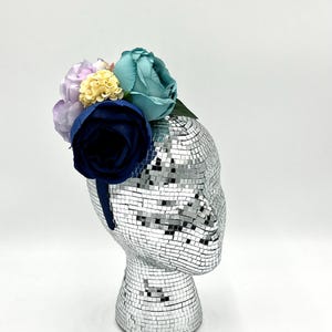May include: A blue, yellow, and purple flower headband with a navy blue velvet band. The headband is on a silver mannequin head.