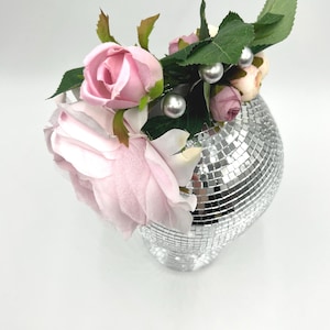 May include: A silver disco ball vase filled with pink roses, smaller pink buds, green leaves, and silver pearl accents. The vase has a heart shape and is covered in small, reflective squares. A decorative floral arrangement.