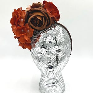 May include: A decorative headpiece with orange and brown artificial flowers. The flowers are attached to a dark headband and sit on a mirrored disco ball head form. The head form is silver and reflects light.