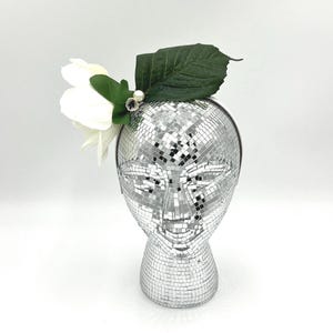 May include: A silver disco ball head with a white flower, green leaves, and a pearl accent. The headpiece is a unique accessory, perfect for themed parties or events. The head is made of small, reflective squares.