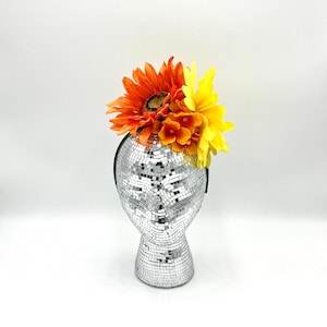 May include: A headband with a cluster of orange and yellow artificial sunflowers. The headband is displayed on a silver mirrored mannequin head.
