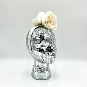 May include: A white headband with two white roses on top. The headband is on a silver mannequin head.