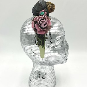 May include: A silver mosaic head sculpture adorned with a floral arrangement. The flowers include a large pink rose, dark gray blooms, and a small brown pinecone. The head is made of small, reflective tiles.