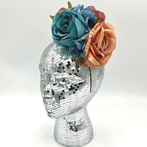May include: A decorative headpiece featuring a silver mirrored head form with a cluster of colorful faux flowers. The flowers include teal roses, blue hydrangeas, and peach-colored roses, creating a vibrant floral arrangement.
