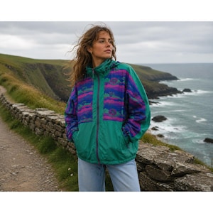 May include: A teal windbreaker jacket featuring a purple and blue patterned panel across the chest and sleeves. The jacket has a full-length front zip, a hood, and long sleeves with elastic cuffs, constructed from a lightweight synthetic material.
