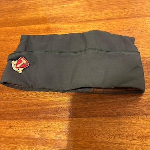 May include: A dark green military garrison cap with a red and white emblem featuring a hammer. The cap is made of a durable fabric and has a brown leather-like interior.