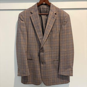 May include: A brown and gray plaid blazer with a two-button closure, notched lapels, and a chest pocket. The blazer is hanging on a wooden hanger against a white wall. The blazer has a classic, tailored fit.