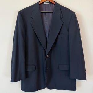 May include: A navy blue blazer with a two-button closure, notched lapels, and two front pockets. The blazer is hanging on a wooden hanger. The interior lining is a darker shade of blue.
