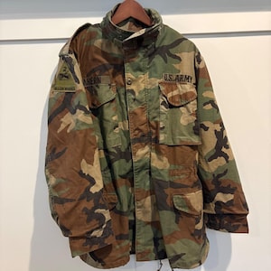 Puede incluir: Chaqueta militar de camuflaje con múltiples bolsillos, cuello alto y las palabras "U.S. ARMY" impresas en la parte delantera. La chaqueta tiene parches en las mangas con el nombre "AHERN" y las palabras "WILL ON WHEELS".