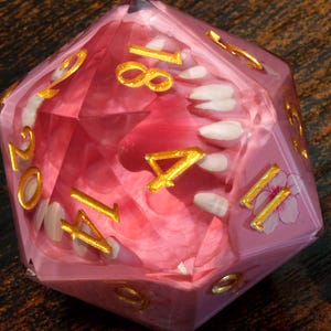 May include: A pink, multi-sided die with gold numbering and white floral accents. The die is made of a translucent material, with a pink floral design visible inside. A unique gaming accessory.