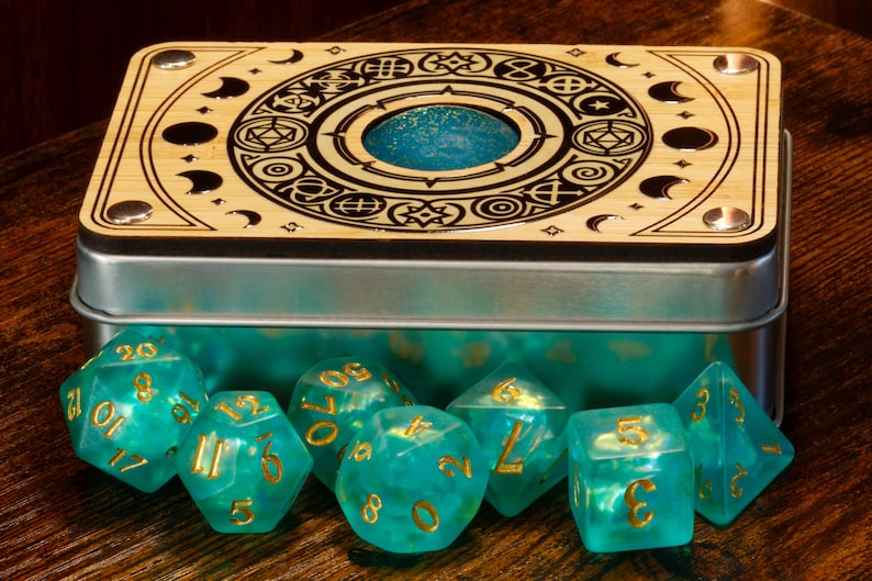 K&ouml;nnte beinhalten: Eine Holzkiste mit einem kreisf&ouml;rmigen Design und einem blau schimmernden Deckel, die tealfarbene, schimmernde W&uuml;rfel mit goldenen Zahlen enth&auml;lt.