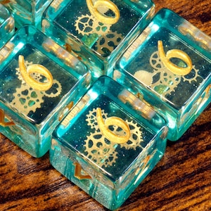 Op de afbeelding: Een set van zes zijdige dobbelstenen van doorschijnende blauwgroene hars met ingebedde goudkleurige tandwielvormige inzetstukken. Elke dobbelsteen toont een opvallend goudkleurig cijfer zes op de bovenkant, met extra tandwieldetails in het materiaal.