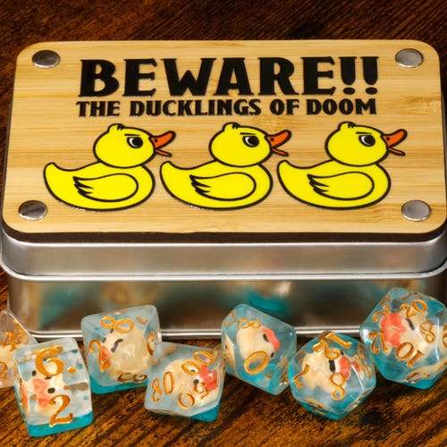 Juego de dados y caja de patitos – ¡Cuidado! ¡Los patitos de la perdición! Dados con un simpático patito de goma en su interior. Accesorio para juegos de rol.