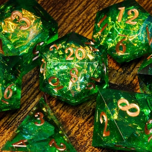 May include: A set of seven polyhedral dice made of translucent green resin with embedded gold-coloured foil flakes. Each die features engraved numbers filled with metallic gold paint, including a twenty-sided die showing the number twenty.