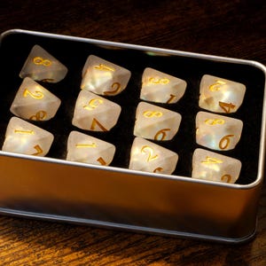 May include: A silver metal box with a black interior holds 16 iridescent white dice with gold numbers. The dice are arranged in four rows of four.