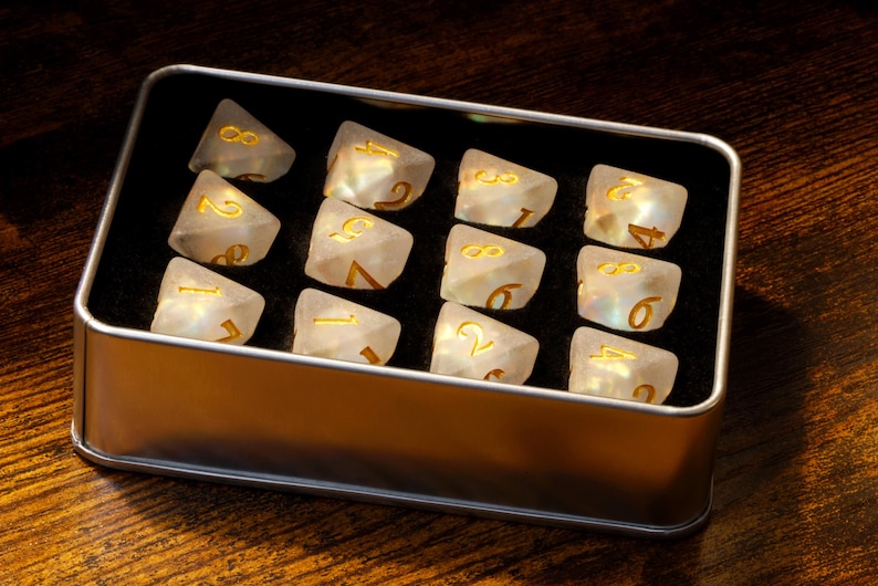 May include: A silver metal box with a black interior holds 16 iridescent white dice with gold numbers. The dice are arranged in four rows of four.