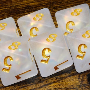 May include: Set of six iridescent white dice with gold numbers. The dice are shaped like cubes and have a pearlescent finish.