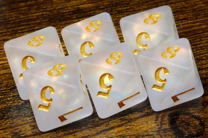 May include: Set of six iridescent white dice with gold numbers. The dice are shaped like cubes and have a pearlescent finish.