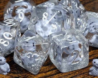 May include: A set of clear polyhedral dice featuring embedded miniature grey figures of small creatures with large ears. Each die contains silver glitter at the base and displays engraved numbers filled with a metallic silver finish.