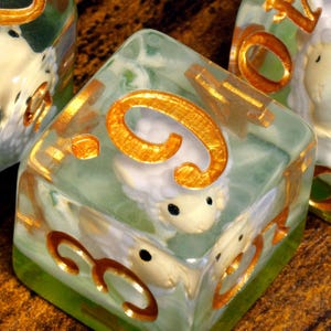 May include: A set of translucent, light green resin dice featuring embedded miniature white sheep figures with black eyes. Each die has large, metallic gold-colored numbers engraved on the exterior surfaces, providing a clear contrast against the pale green and white interior elements.