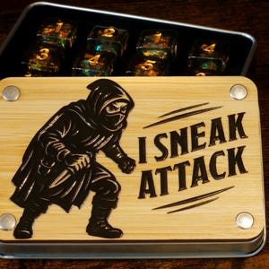 Op de afbeelding: Een houten doos met een metalen frame, met een ninja-afbeelding en de tekst "I SNEAK ATTACK". Binnenin bevinden zich verschillende doorzichtige dobbelstenen met goudkleurige cijfers. De doos staat op een donker houten oppervlak.