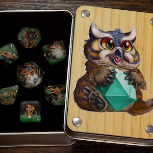 May include: A rectangular metal storage box with a bamboo lid featuring a colourful illustration of an owl holding a green geometric shape. The open box contains a set of seven translucent green polyhedral dice with gold numbers.