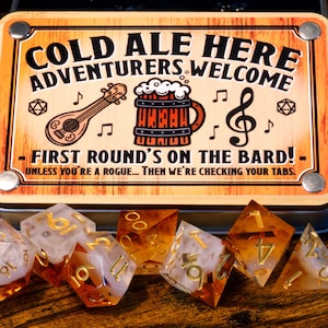 May include: A wooden sign with the text "COLD ALE HERE ADVENTURERS WELCOME" and the phrase "FIRST ROUND'S ON THE BARD!" is displayed above a set of polyhedral dice. The dice are clear with gold numbers and amber-colored accents.