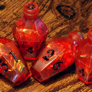 May include: Four faceted, red, potion-shaped dice with black numbers. The dice have a glittery, translucent appearance. The tops of two dice have a black starburst design. The dice are arranged on a wooden surface.