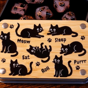 Könnte beinhalten: Eine Holzkiste mit einem schwarzen Katzenmotiv. Die Kiste hat einen Deckel und ist geöffnet, um einen Satz Würfel mit schwarzen Katzen darauf zu enthüllen. Die Würfel sind in verschiedenen Farben, darunter Schwarz, Weiß und Gold. Die Kiste hat die Wörter "Meow", "Eat", "Sleep", "Purrr" und "Roll" darauf gedruckt.